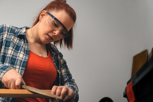 close-up of a of a young red-haired carpenter woman, concentrated and precise, working on the design of wood in a small carpentry workshop, dressed in a blue checked shirt and a red t-shirt. Carpenter woman holding a nail in a wooden board, in her small carpentry business. Warm light indoors, background with wooden slats. Horizontal.