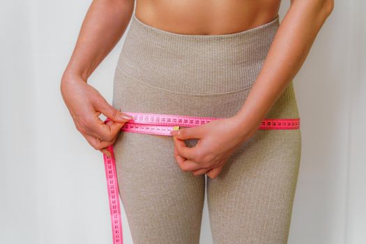 Cropped view of slim woman measuring hips with tape measure at home, close up. An unrecognizable European woman checks the result of a weight loss diet or liposuction indoors. Healthy lifestyle