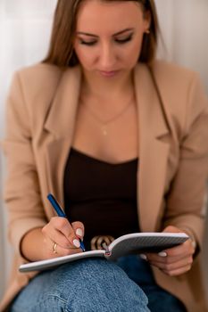 European woman writes in a notebook. She is wearing a beige jacket and jeans