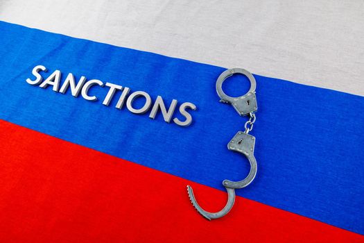 the word sanctions laid with silver metal letters on russian tricolor flag near handcuffs over it in directly above view
