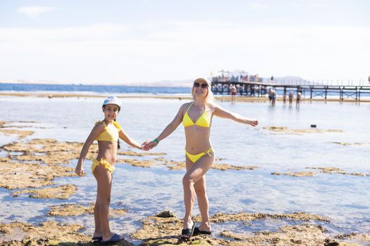 Smiling mother and beautiful daughter having fun on the beach. Portrait of happy woman giving a piggyback ride to cute little girl with copy space.