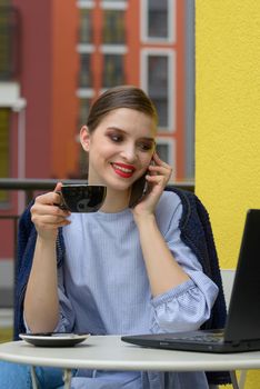 Charming happy woman student communicates by phone and use laptop computer to prepare for the course work. Concept of working outdoors