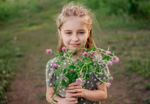 Beautiful preteen girl with incredible eyes looking at the camera and holding flower bouquet on the nature. Portrait of cute female kid with hairstyle in the field in summer time