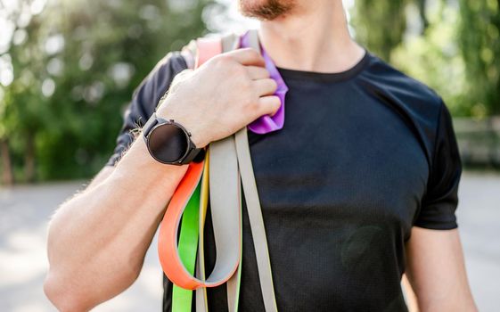 Sportive man guy holding in his hand wearing fitness tracker set of colorful elastic rubber bands at the stadium outdoors. Athlete male person during workout with additional sport equipmant