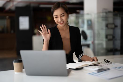 Young asian businesswoman making video call on laptop at home office, waving at screen, chatting with, clients, colleagues. Working remotely, freelance, online lecturer, teacher.
