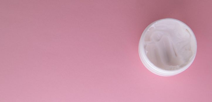White cosmetic face cream in a round open jar on a pink background, top view, copy of the space on the left.