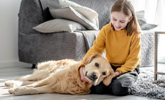 Preteen girl playing with golden retriever dog sitting on floor. Pretty kid child petting doggy pet at home