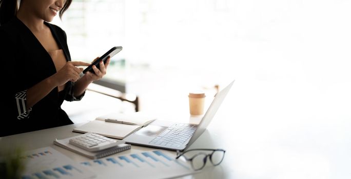 Asian woman holding smart phone while using calculator and laptop for business financial accounting calculate money bank loan rent payments manage expenses finances taxes doing paperwork concept, close up