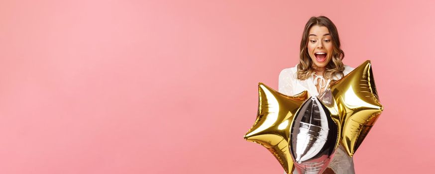 Holidays, celebration and women concept. Portrait of happy lovely young woman in white dress, gasping from amazement and joy, holding birthday star-shaped balloons, pink background.