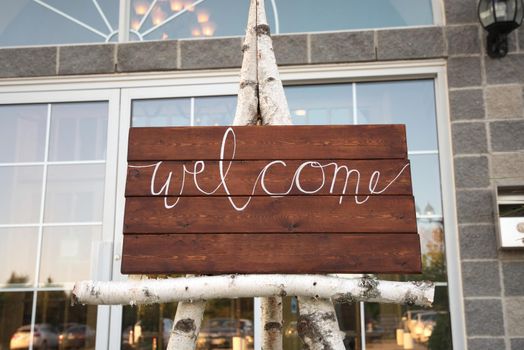Rustic Handmade Wood Welcome Sign with Room for Text Outside of Fancy Venue with Elegant Windows. Displayed on Birch Log Easel