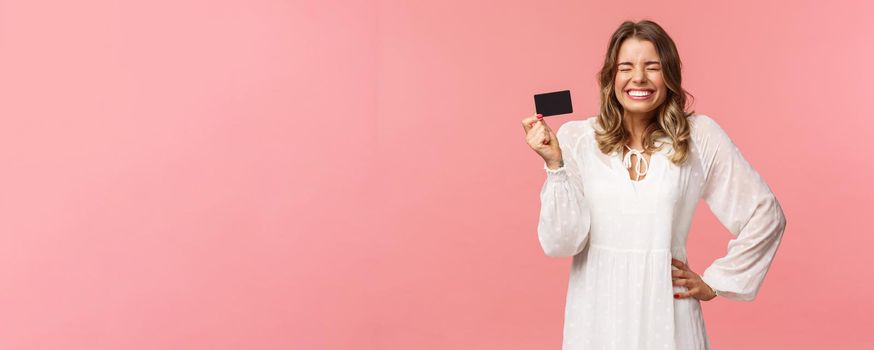 Portrait of excited cute and silly, feminine blond girl in white dress, close eyes giggle and smiling happy, got her first payment new job, holding credit card, use banking service, pink background.