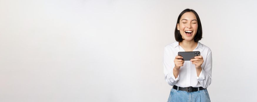 Image of young asian woman watching on smartphone app, holding mobile phone and looking at screen, laughing and smiling, standing over white background.