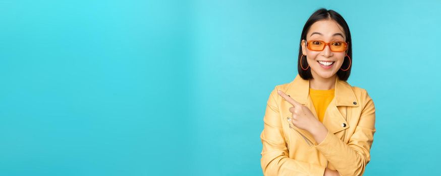 Stylish smiling asian woman in sunglasses, pointing finger left, showing advertisement banner, standing in trendy yellow coat over blue background.