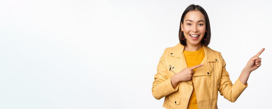 Enthusiastic asian girl pointing fingers right, showing advertisement and smiling, inviting to the store, standing over white background.