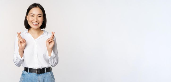 Portrait of young korean woman, asian girl cross fingers and praying, making wish, anticipating, waiting for results, standing over white background.