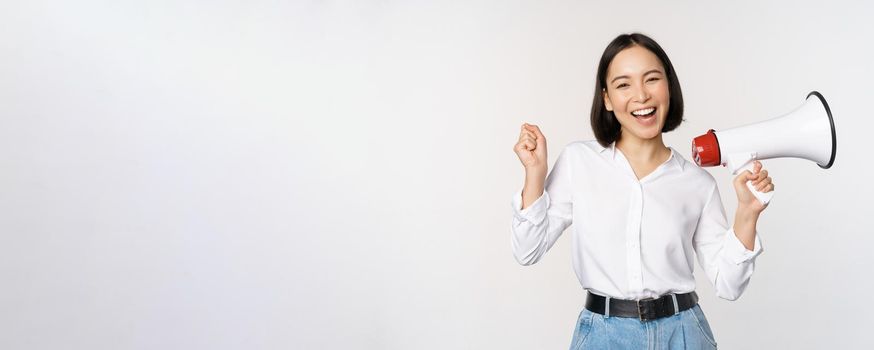 Smiling young asian woman posing with megaphone, concept of news, announcement and information, standing over white background.