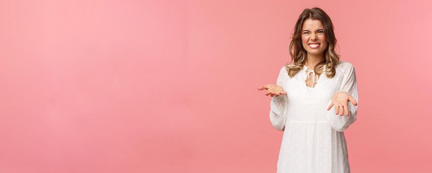 Portrait of dissatisfied, disappointed cute girl in white dress, cringe show tongue, smell something awful, pointing with raised hands at bad unappropriate thing, grimacing dislike, pink background.