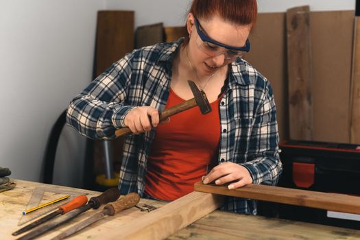 Detail of the hands of a young red-haired carpenter woman, concentrated and precise, working on the design of the wood in a small carpentry workshop, dressed in a blue checked shirt and a red t-shirt. Woman carpenter hammering a nail into a wooden board in her small carpentry workshop. Warm light indoors, background with wooden slats. Horizontal.