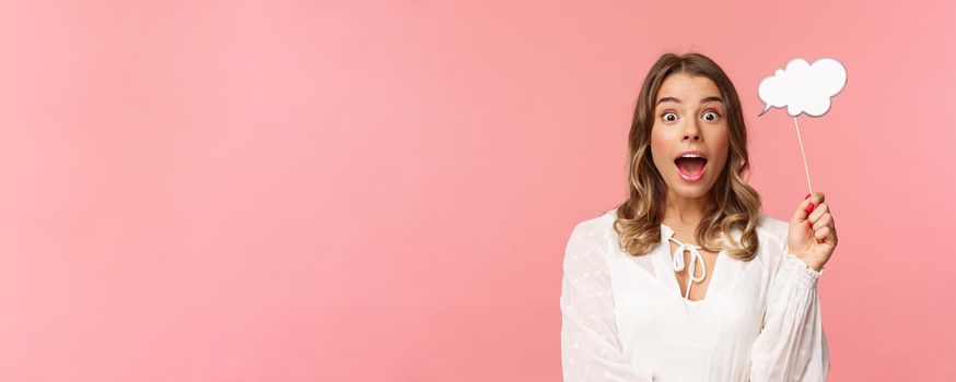 Spring, happiness and celebration concept. Close-up portrait of surprised, excited blond girl looking amazed, holding cloud comment stick near head and gasping astonished, have idea, pink background.