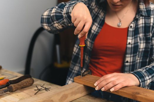 Close up shot of the hands of a young red-haired woman carpenter, concentrated and precise, working on the layout of wood in a small carpentry workshop, dressed in a blue checked shirt and a red t-shirt. Young woman carpenter using screwdriver on wooden plank. Warm light indoors, background with wooden slats. Horizontal.