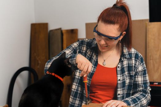 Close-up of the hands of a young red-haired carpenter, concentrated and precise, working on the layout of wood in a small carpentry workshop, dressed in a blue chequered shirt and a red T-shirt. Female carpenter in the company of her pet black furred cat in her carpentry workshop. Warm light indoors, background with wooden slats. Horizontal.