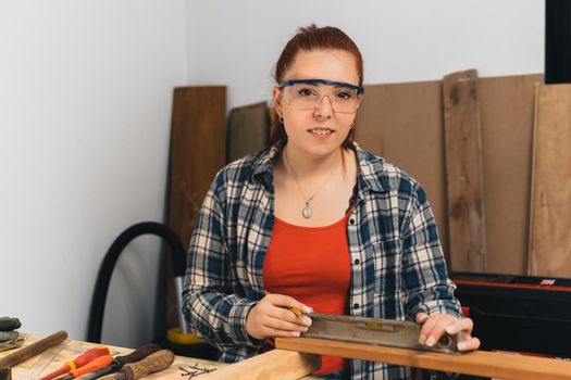 Close-up of the hands of a young red-haired carpenter, concentrated and precise, working on the layout of timbers in a small carpentry workshop, dressed in a blue checked shirt and a red T-shirt. Female carpenter using a precision level in her small carpentry workshop. Warm light indoors, background with wooden slats. Horizontal. Looking at the camera