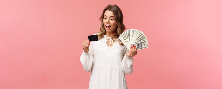 Portrait of rich good-looking blond girl in white dress, holding dollars money and credit card, winning lottery, think how invest, standing happy pink background. Copy space