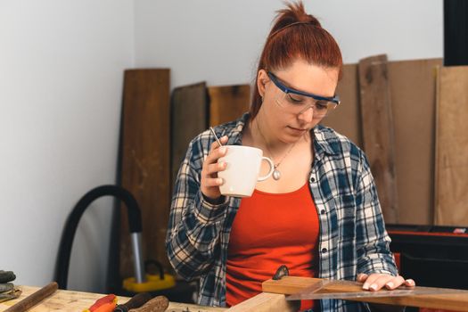 Close-up of the hands of a young red-haired carpenter, concentrated and precise, working on the layout of timbers in a small carpentry workshop, dressed in a blue checked shirt and a red T-shirt. Woman carpenter having a coffee, planning her new project in her small carpentry business. Warm light indoors, background with wooden slats. Horizontal.