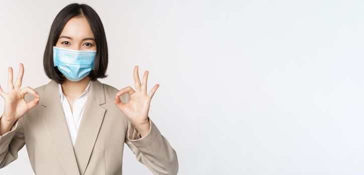 Coronavirus and workplace concept. Image of asian saleswoman, company worker in medical mask showing okay sign, smiling pleased, white background.