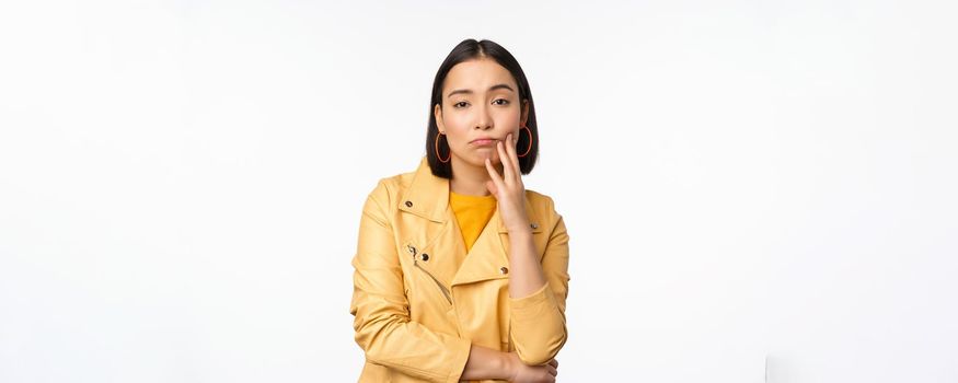 Image of stylish japanese girl looking with unamused, tired and upset face expression, standing in casual clothes over white background. Copy space