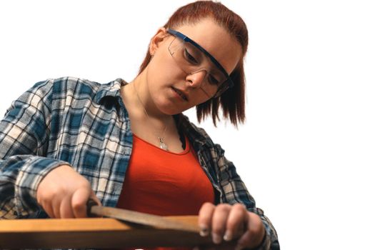 close-up of a of a young red-haired carpenter woman, concentrated and precise, working on the design of wood in a small carpentry workshop, dressed in a blue checked shirt and a red t-shirt. Carpenter woman holding a nail in a wooden board, in her small carpentry business. Warm light indoors, white background. Horizontal.