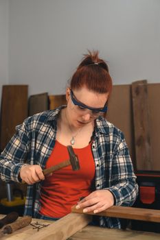 Detail of the hands of a young red-haired carpenter woman, concentrated and precise, working on the design of wood in a small carpentry workshop, dressed in a blue checked shirt and a red t-shirt. Carpenter woman holding a nail in a wooden board, in her small carpentry business. Warm light indoors, background with wooden slats. Vertical