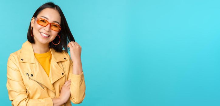 Young stylish asian woman in sunglasses smiling, playing with her haircut and looking happy, posing against blue background.