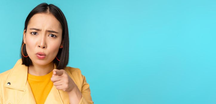 Close up of asian girl accusing and pointing finger at camera, looking offended or displeased, scolding you, standing over blue background.