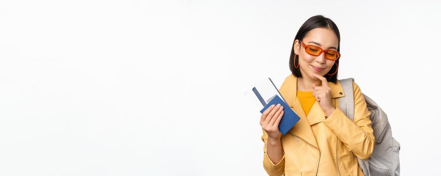 Asian girl tourist with boarding tickets and passport going abroad, holding backpack, thinking of travelling, standing over white background.