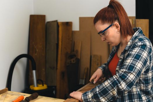 young red-haired carpenter woman, concentrated and precise, working on the design of wood in a small carpentry workshop, dressed in a blue checked shirt and a red t-shirt. Carpenter woman holding a nail in a wooden board, in her small carpentry business. Warm light indoors, background with wooden slats. Horizontal.