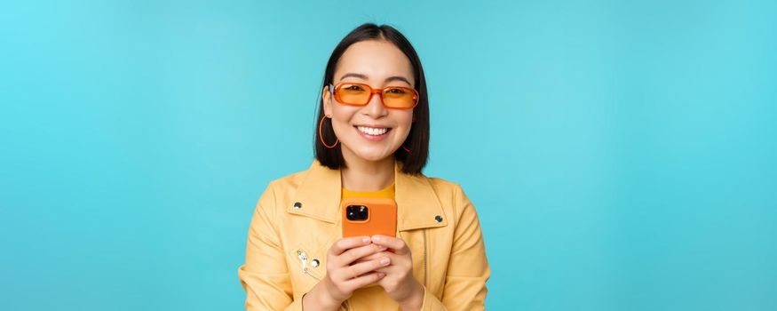 Portrait of enthusiastic asian woman in sunglasses, using mobile phone, smiling and laughing, looking happy, holding smartphone, standing over blue background.