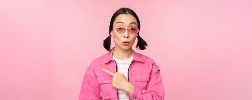 Close up portrait of asian woman gasping, looking surprised, pointing finger at banner, advertisement, standing over pink background.