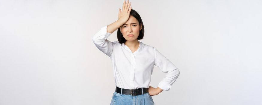 Bothered and tired asian woman, looking complicated, slap forehead, facepalm sign and grimacing upset, looking distressed at camera, white background.
