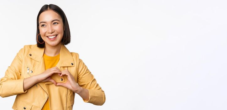 Image of beautiful asian girl showing heart, love sign, express care and romantic feelings, standing over white studio background.