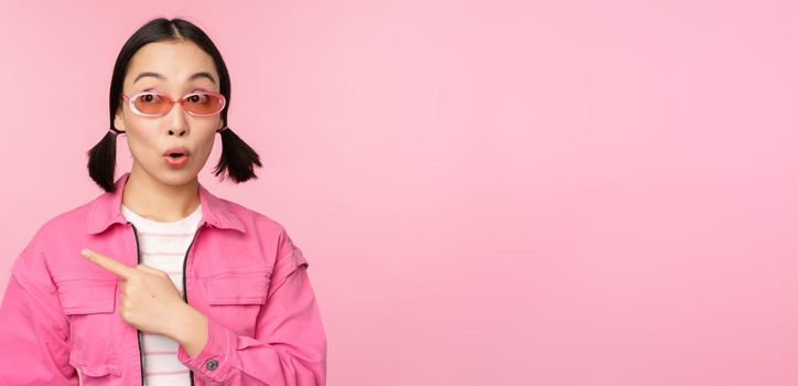 Portrait of smiling asian girl in stylish outfit, sunglasses, pointing finger left, showing advertisement, banner, standing over pink background. Copy space