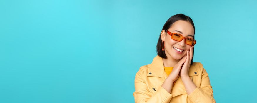 Close up portrait of asian young woman in sunglasses, smiling and looking romantic, standing happy over blue background. Copy space