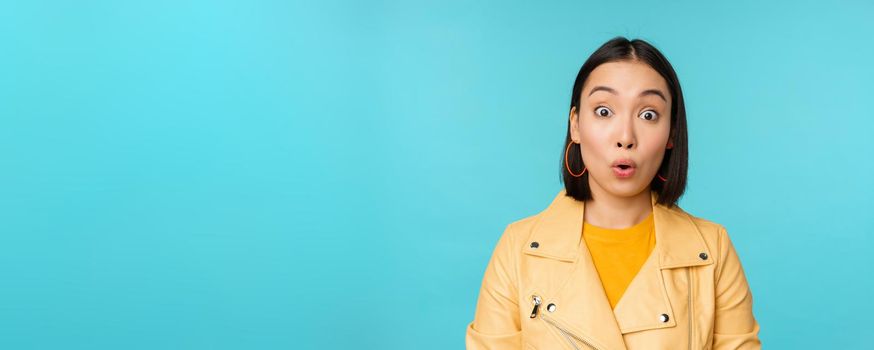 Close up portrait of asian girl looks surprised at amazed, wow face expression, stands over blue background.