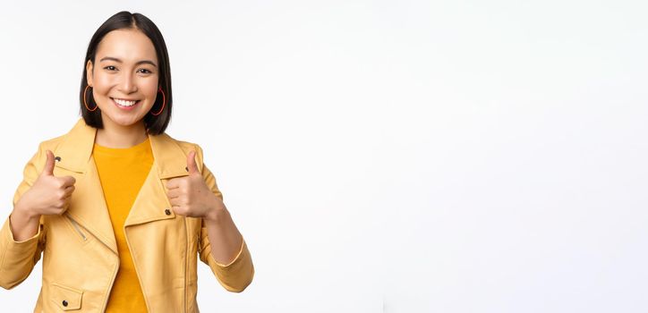 Cheerful asian woman smiling pleased, showing thumbs up in approval, standing in casual clothes over white background.