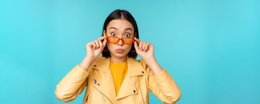 Close up portrait of asian girl in sunglasses, looking happy and surprised, excitement on face, standing over blue background.