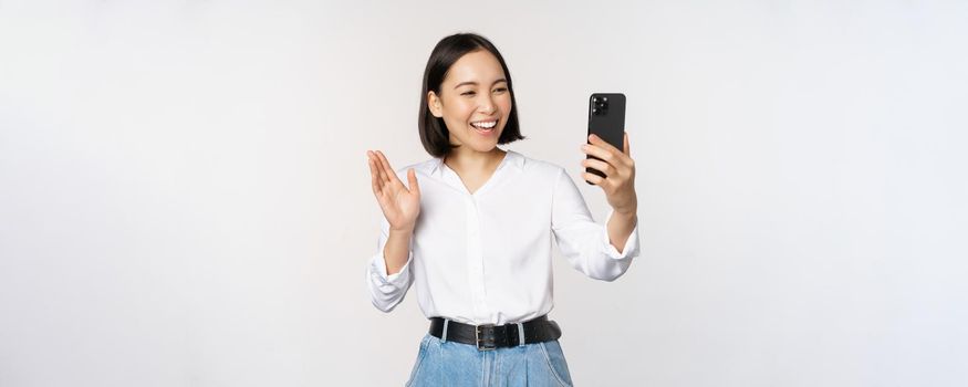 Image of stylish modern asian woman waving hand at smartphone front camera, video chat, chatting with person on mobile phone application, white background.