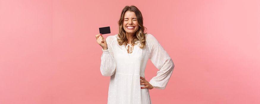 Portrait of excited cute and silly, feminine blond girl in white dress, close eyes giggle and smiling happy, got her first payment new job, holding credit card, use banking service, pink background.