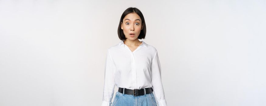 Portrait of surprised young office woman, asian businesswoman gasping amazed, saying wow, standing impressed of news against white background.