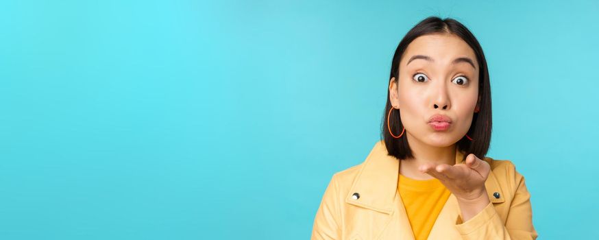 Close up portrait of funny asian girl sending air kiss, blowing at camera with popped eyes, standing over blue background.