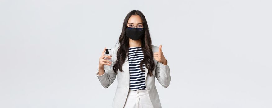 Business, finance and employment, covid-19 preventing virus and social distancing concept. Smiling businesswoman in face mask showing hand sanitizer and thumbs-up, ask to use it.